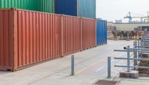 Contenedores de transporte marítimo apilados en un puerto, con ganado bovino en un corral cercano y una grúa portuaria al fondo.