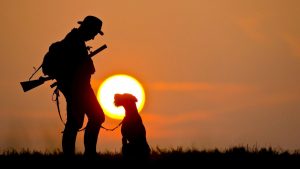 Silueta a contraluz de un cazador con una escopeta al hombro junto a su perro sentado, durante el atardecer